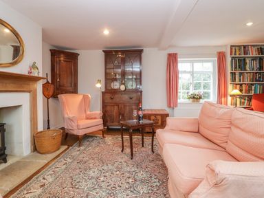 A living room with pink upholstered sofa and armchair a wooden cabinet a small wooden table with wine on a rug near a window with pink curtains at Church View in Salisbury