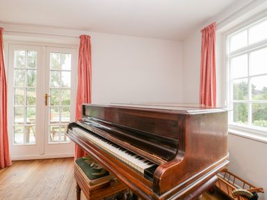 A wooden piano with a bench near French doors and windows with red curtains at Church View in Salisbury