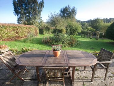 A wooden outdoor table with three chairs and a potted plant on a patio facing a green garden with trees and hedges at Church View in Salisbury