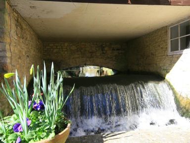 A stone bridge over flowing water with flowers in a pot in the foreground at Cider Press in Burford
