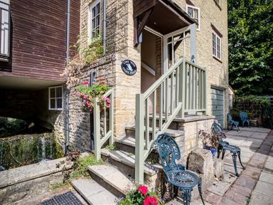An outdoor stone staircase with a green railing leading to an open door on a stone building with metal chairs and potted flowers at Cider Press in Burford