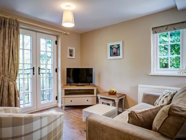 A living room with a sofa, armchair, TV on a stand, side tables, and large windows at Cider Press in Burford