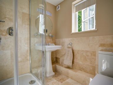 A bathroom with a glass shower enclosure a pedestal sink a round mirror a towel ring with a towel a window and a toilet at Cider Press in Burford
