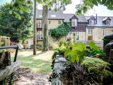 An outdoor garden area with grass trees and plants in front of a stone house with balconies and windows at Cider Press in Burford
