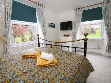 A bedroom with a patterned bedspread and towels on the bed two windows with blinds and curtains a chest of drawers and a wall mounted television at Giddy Goose in Windermere