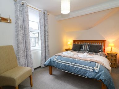 A bedroom with a double bed with pillows and bedside tables with lamps by a window with curtains and a chair at Giddy Goose in Windermere