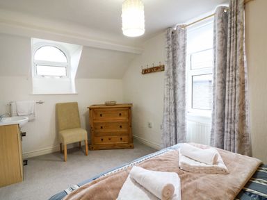 A bedroom with a bed, folded towels, a wooden chest of drawers, a chair, a small sink, and two windows with curtains at Giddy Goose in Windermere