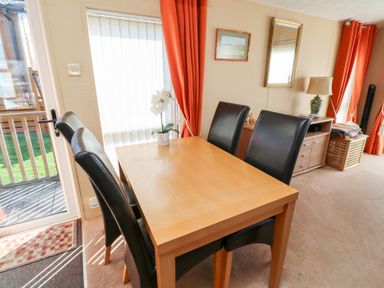 A dining area with a wooden table and four black chairs next to a glass door with vertical blinds orange curtains and a sideboard with a lamp at Percy Wood Country Park in Swarland