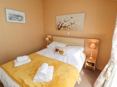 A bedroom with a double bed covered with white and yellow bedding two bedside tables with lamps artwork on the walls and patterned curtains at Percy Wood Country Park in Swarland