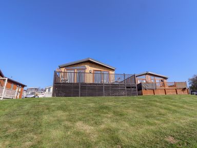 A wooden lodge with a deck on a grassy hill under a clear blue sky at Percy Wood Country Park in Swarland