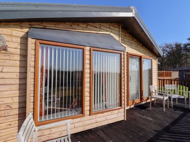 Wooden cabin exterior with large windows and vertical blinds on a deck with wooden chairs at Percy Wood Country Park in Swarland