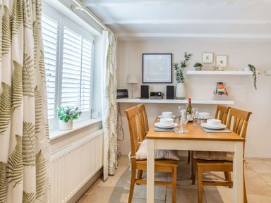 A dining area with a wooden table and chairs set with plates glasses and a wine bottle near a window with curtains and shelves with plants and decorations at Beach Retreat in Instow