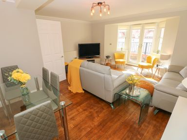 A living room with grey sofas yellow chairs a glass dining table and flowers at Seashells in Tenby