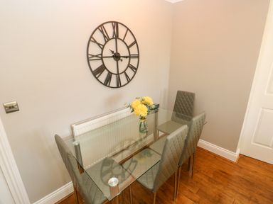 A dining area with a glass table and four gray chairs a large wall clock and a vase with yellow flowers at Seashells in Tenby