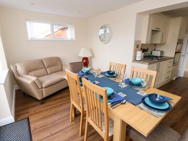 A dining area with a wooden table set for six next to a beige sofa and a kitchen at Elliotts Retreat in Sheringham