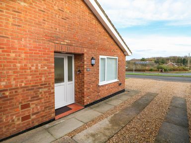 A brick house entrance with a white door and window beside a gravel driveway at Elliotts Retreat in Sheringham