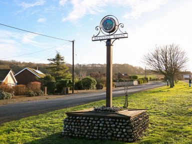 A village sign on a stone base beside a road with houses trees and grassy area at Elliots Retreat in Sheringham