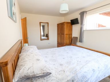 A bedroom with a wooden bed frame a floral bedspread a wooden wardrobe a wall mirror a chair and a window with gray curtains at Elliotts Retreat in Sheringham