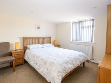 A bedroom with a double bed two bedside tables lamps a chair a window with vertical blinds and wooden furniture at Elliotts Retreat in Sheringham