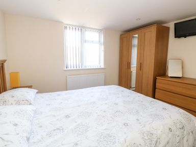 A bedroom with a bed and floral bedding a wooden wardrobe with a mirror a dresser with a mirror and a wall-mounted TV at Elliotts Retreat in Sheringham