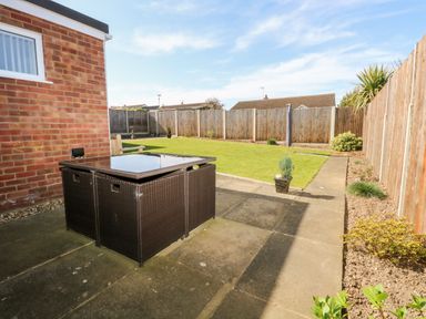 A backyard with a paved patio area a wicker table a grassy lawn and wooden fencing at Elliotts Retreat in Sheringham