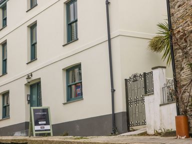 The exterior of a white building with green window frames a sign board on the sidewalk and a black wrought iron gate at Lantern Cottage in Padstow