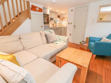 A living room with beige sofas a wooden coffee table a blue armchair and a kitchen area at Lantern Cottage in Padstow