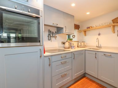 A kitchen with blue cabinets a built-in oven countertop appliances utensils on a wall rack and a wooden shelf with cups at Lantern Cottage in Padstow