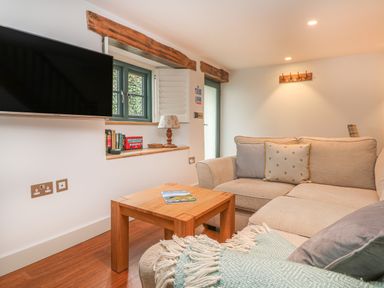 A living room with a beige sofa, wooden coffee table, wall-mounted TV, and a window with wooden beams at Lantern Cottage in Padstow