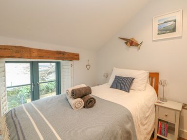 A bedroom with a bed with folded towels a bedside table with a lamp and books and a window with wooden shutters at Lantern Cottage in Padstow