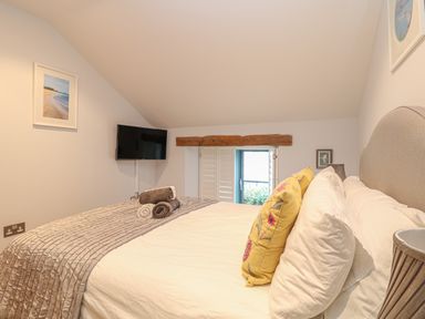 A bedroom with a bed with white bedding and yellow pillows towels on the bed a TV mounted on the wall and a small window with wooden shutters at Lantern Cottage Padstow