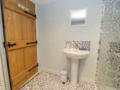 A bathroom with a wooden door a pedestal sink a small mirror a trash bin and patterned tiles on floor and shower at Wagtail Nest Northfield Cottage Scalby