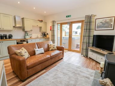 A living room with a brown leather sofa kitchen area and tv near glass doors with curtains at Wagtail Nest Northfield Cottage in Scalby