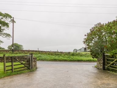 A driveway with open wooden gates leading to a road and green fields with stone walls at Wagtail Nest Northfield Cottage Scalby