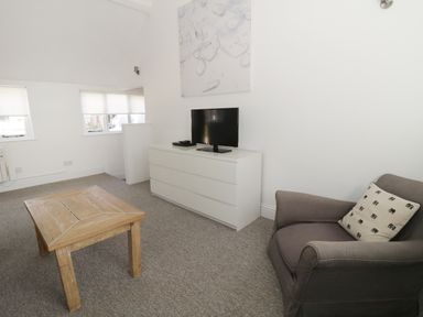 A living room with a wooden coffee table a gray armchair with a patterned pillow a white dresser with a television on top and three windows with blinds at Orchard Cottage in Shaftesbury