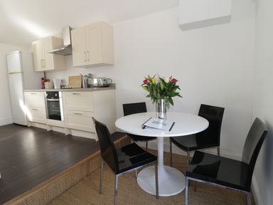 A kitchen area with cream cabinets and a white refrigerator next to a round white table with a vase of flowers and black chairs at Orchard Cottage in Shaftesbury