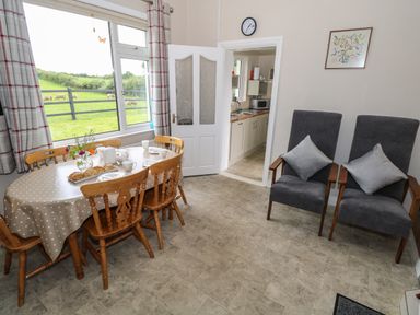 A dining area with a table set for tea wooden chairs two gray armchairs and a kitchen visible through a door at Kates Cottage near Ballina County Mayo