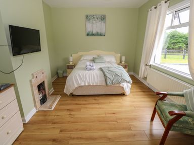 A bedroom with a bed nightstands lamps a wall mounted television a small fireplace a green armchair and a large window at Kates Cottage near Ballina County Mayo