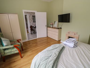 A bedroom with a bed and folded towels a green armchair a chest of drawers with flowers a wall-mounted TV and an open door at Kates Cottage near Ballina County Mayo