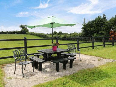 An outdoor picnic area with a black table and benches two metal chairs and a green umbrella on a concrete slab in a grassy fenced yard at Kates Cottage near Ballina County Mayo