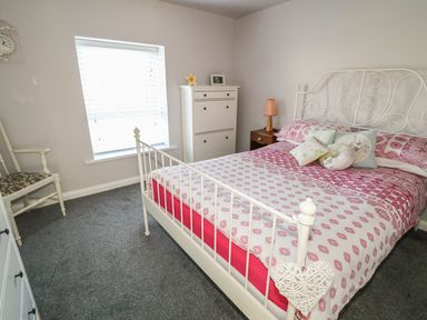 A bedroom with a bed with red and white bedding a chair a window with blinds a white cabinet and a bedside table with a lamp at Cranny Corner in Carnlough