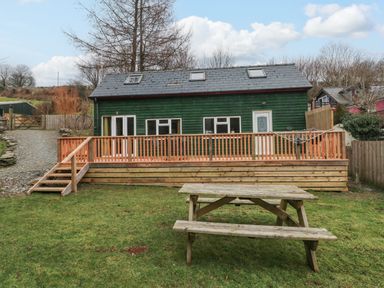 A backyard with a wooden picnic table a deck with railings and steps and a green wooden house with windows and a door at The Hen Pen in Star near Cenarth