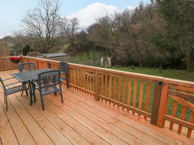 A wooden deck with a metal table and four chairs and a barbecue grill overlooking a grassy yard with trees at The Hen Pen Star near Cenarth