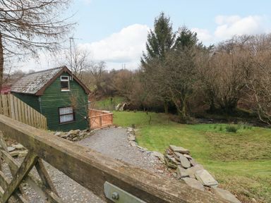 A small green wooden house with a gravel path and grassy area surrounded by trees at The Hen Pen in Star near Cenarth