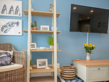 A living area with blue walls featuring a wicker chair with cushions a wooden shelf with plants and framed pictures a wicker basket a TV mounted on the wall and a wooden coffee table with a vase of yellow flowers at Coastal Haven in Crackington near Crackington Haven