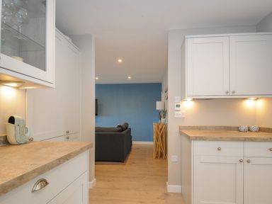 A kitchen with white cabinets and beige countertops leading to a living room with a blue wall and a dark grey sofa at Coastal Haven in Crackington near Crackington Haven