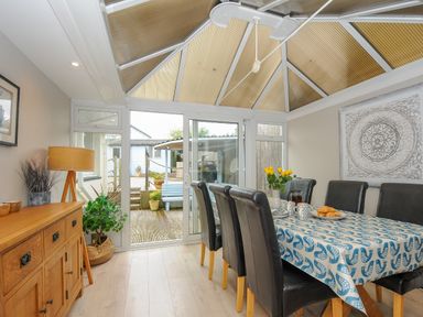 A dining room with a table covered with a patterned tablecloth chairs and a sideboard with a lamp and plants at Coastal Haven in Crackington near Crackington Haven