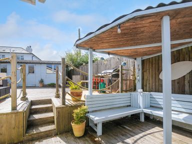 An outdoor seating area with a wooden bench and roof near a wooden deck with stairs and a barbecue at Coastal Haven in Crackington near Crackington Haven