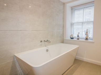 A bathroom with a rectangular bathtub and a window with blinds and decorative items at Midships in Mevagissey