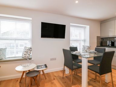 A dining area with a glass table and four chairs near windows with blinds and cushions at Midships in Mevagissey